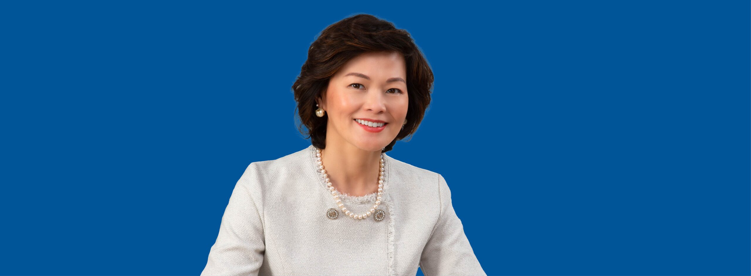 Elegant woman smiling, pearl necklace, white blazer, blue background.