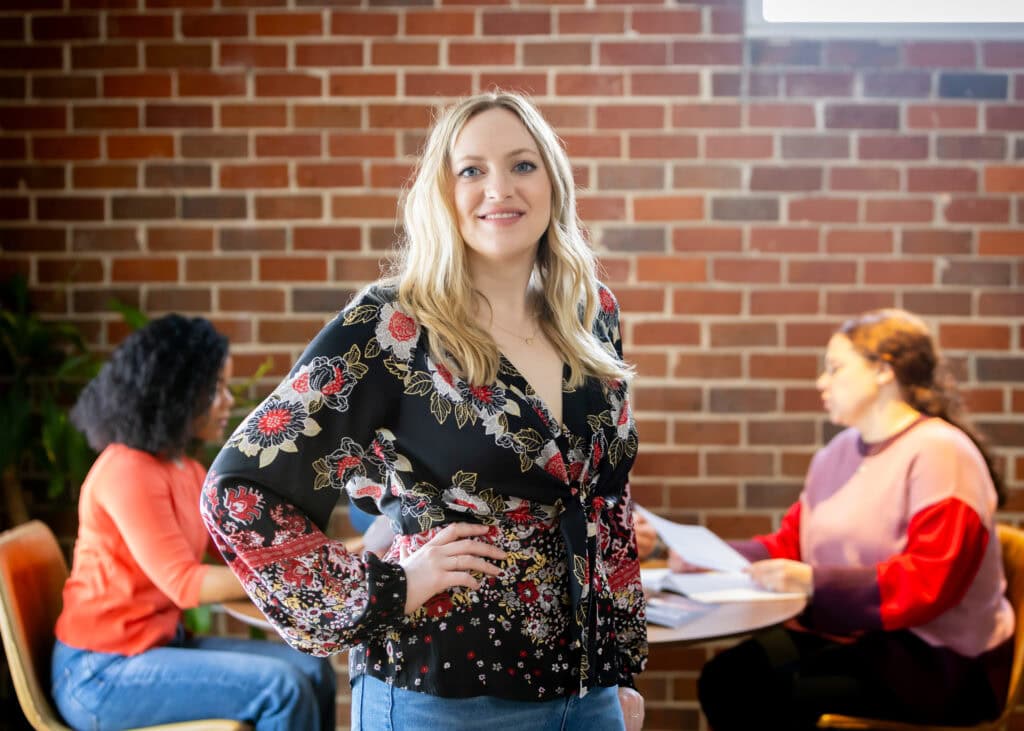 Woman in floral top standing in office, colleagues behind.