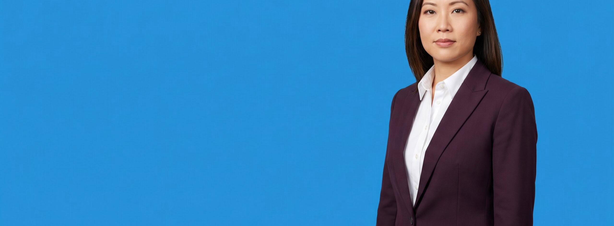 Asian woman professional in her late 30s, average build, dark plum blazer and white shirt, on a blue studio background — Criminal Defense Law Firm Marketing: How to Reach Clients Who Need You Right Now.